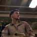 A young male individual wearing USMC MARPAT desert camouflage combat shirt is seated in what appears to be an indoor military or industrial environment. Tactical gear and equipment are visible in the foreground, and a weapon system is partially visible to the left. The subject is looking off to one side under artificial overhead lighting.