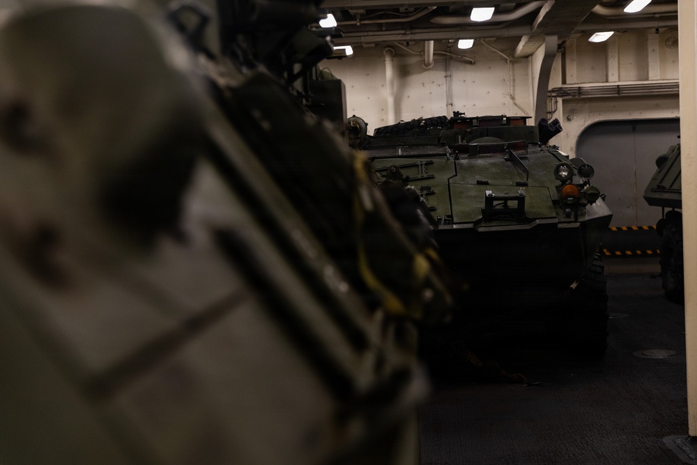 The image shows military armored vehicles stored inside what appears to be the well deck or vehicle bay of a naval amphibious assault ship. At least two armored personnel carriers, likely AAV7 Assault Amphibious Vehicles, are visible parked in the enclosed space. The interior features white-painted bulkheads, overhead lighting, and safety markings typical of a naval vessel.