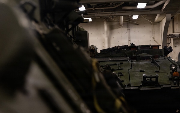 The image shows military armored vehicles stored inside what appears to be the well deck or vehicle bay of a naval amphibious assault ship. At least two armored personnel carriers, likely AAV7 Assault Amphibious Vehicles, are visible parked in the enclosed space. The interior features white-painted bulkheads, overhead lighting, and safety markings typical of a naval vessel.