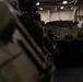 The image shows military armored vehicles stored inside what appears to be the well deck or vehicle bay of a naval amphibious assault ship. At least two armored personnel carriers, likely AAV7 Assault Amphibious Vehicles, are visible parked in the enclosed space. The interior features white-painted bulkheads, overhead lighting, and safety markings typical of a naval vessel.