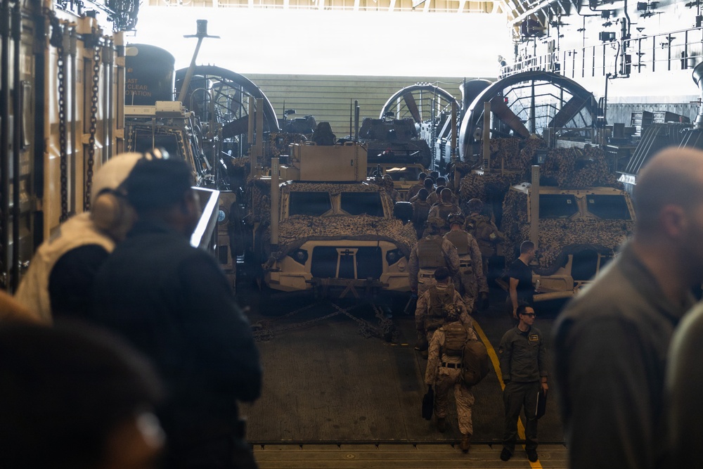 The image shows the interior of a military vessel's well deck or cargo bay, loaded with camouflage-painted tactical military vehicles, likely MRAPs or Joint Light Tactical Vehicles (JLTVs). Military personnel in uniform are walking through the vehicle bay, suggesting a loading, unloading, or inspection operation. Large mechanical winch or pulley systems are visible along the sides and rear of the bay.