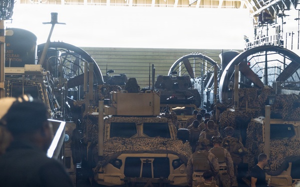 The image shows the interior of a military vessel's well deck or cargo bay, loaded with camouflage-painted tactical military vehicles, likely MRAPs or Joint Light Tactical Vehicles (JLTVs). Military personnel in uniform are walking through the vehicle bay, suggesting a loading, unloading, or inspection operation. Large mechanical winch or pulley systems are visible along the sides and rear of the bay.