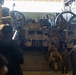 The image shows the interior of a military vessel's well deck or cargo bay, loaded with camouflage-painted tactical military vehicles, likely MRAPs or Joint Light Tactical Vehicles (JLTVs). Military personnel in uniform are walking through the vehicle bay, suggesting a loading, unloading, or inspection operation. Large mechanical winch or pulley systems are visible along the sides and rear of the bay.