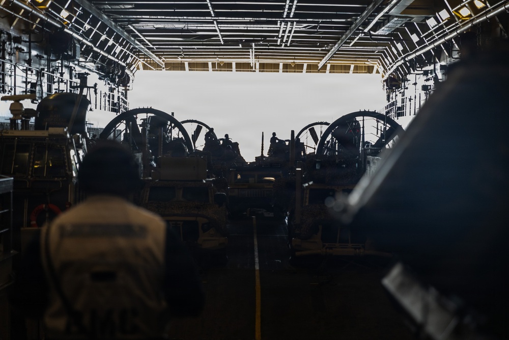 The image shows the interior of a large military vessel's well deck or vehicle bay, viewed from inside looking toward an open stern ramp. Multiple military vehicles and personnel are visible in silhouette against the bright exterior light. The scene appears to depict loading or staging operations of military equipment aboard a naval amphibious ship.