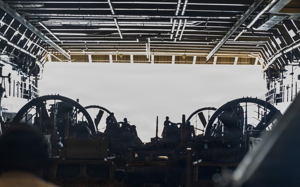 The image shows the interior of a large military vessel's well deck or vehicle bay, viewed from inside looking toward an open stern ramp. Multiple military vehicles and personnel are visible in silhouette against the bright exterior light. The scene appears to depict loading or staging operations of military equipment aboard a naval amphibious ship.