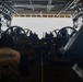 The image shows the interior of a large military vessel's well deck or vehicle bay, viewed from inside looking toward an open stern ramp. Multiple military vehicles and personnel are visible in silhouette against the bright exterior light. The scene appears to depict loading or staging operations of military equipment aboard a naval amphibious ship.