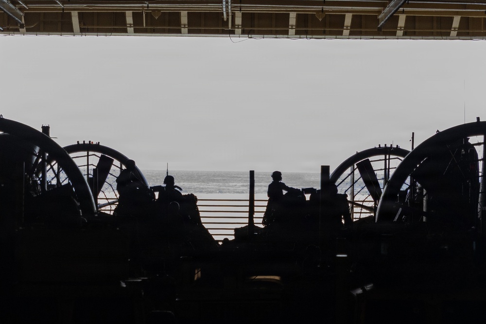 The image shows the interior of what appears to be a naval vessel's well deck or hangar bay, viewed from inside looking out toward the open ocean. Two large circular fan/propeller shrouds are visible on either side, characteristic of Landing Craft Air Cushion (LCAC) hovercraft. Several silhouetted personnel are visible against the bright ocean backdrop, suggesting active operations or standby status.