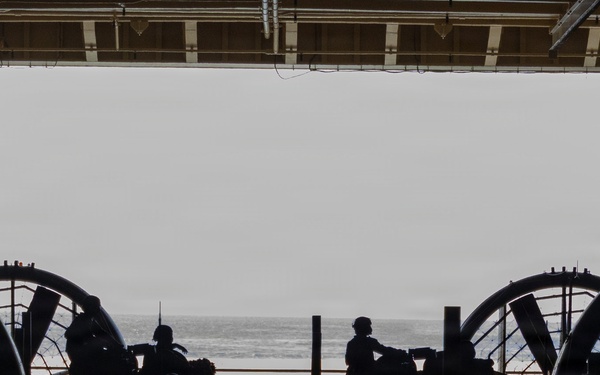 The image shows the interior of what appears to be a naval vessel's well deck or hangar bay, viewed from inside looking out toward the open ocean. Two large circular fan/propeller shrouds are visible on either side, characteristic of Landing Craft Air Cushion (LCAC) hovercraft. Several silhouetted personnel are visible against the bright ocean backdrop, suggesting active operations or standby status.