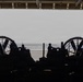 The image shows the interior of what appears to be a naval vessel's well deck or hangar bay, viewed from inside looking out toward the open ocean. Two large circular fan/propeller shrouds are visible on either side, characteristic of Landing Craft Air Cushion (LCAC) hovercraft. Several silhouetted personnel are visible against the bright ocean backdrop, suggesting active operations or standby status.