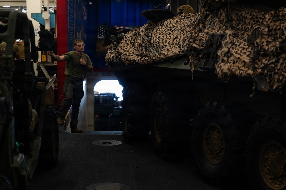 A military service member in a green t-shirt stands in a vehicle storage bay, likely aboard a naval vessel or military transport facility. A large armored vehicle covered with desert camouflage netting dominates the right side of the frame. The space appears to be a well-equipped vehicle deck with multiple military assets stored in close proximity.