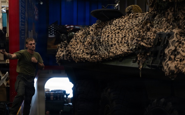 A military service member in a green t-shirt stands in a vehicle storage bay, likely aboard a naval vessel or military transport facility. A large armored vehicle covered with desert camouflage netting dominates the right side of the frame. The space appears to be a well-equipped vehicle deck with multiple military assets stored in close proximity.