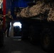 A military service member in a green t-shirt stands in a vehicle storage bay, likely aboard a naval vessel or military transport facility. A large armored vehicle covered with desert camouflage netting dominates the right side of the frame. The space appears to be a well-equipped vehicle deck with multiple military assets stored in close proximity.