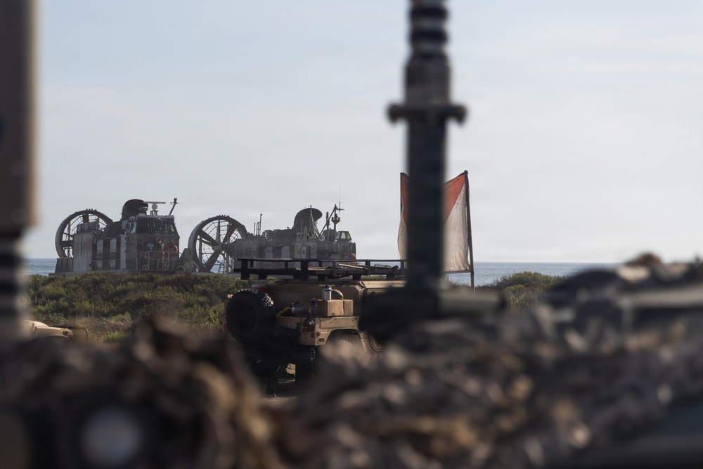 The image shows a military beach landing operation with a Landing Craft Air Cushion (LCAC) hovercraft visible in the background near the shoreline. In the foreground and midground, military ground vehicles are positioned on the beach, with a red and white signal flag visible. The photo is taken from a low angle through or around military equipment, giving a field-level perspective.