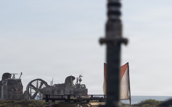 The image shows a military beach landing operation with a Landing Craft Air Cushion (LCAC) hovercraft visible in the background near the shoreline. In the foreground and midground, military ground vehicles are positioned on the beach, with a red and white signal flag visible. The photo is taken from a low angle through or around military equipment, giving a field-level perspective.