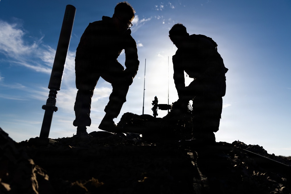 Two individuals are silhouetted against a bright blue sky, working on or near rocky terrain. They appear to be handling equipment, with a cylindrical pole or tube visible on the left side and additional equipment structures in the background. The strong backlit composition creates dramatic dark silhouettes with minimal detail visible on the subjects.