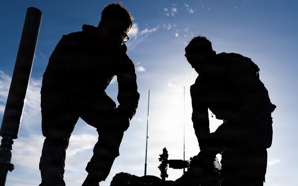 Two individuals are silhouetted against a bright blue sky, working on or near rocky terrain. They appear to be handling equipment, with a cylindrical pole or tube visible on the left side and additional equipment structures in the background. The strong backlit composition creates dramatic dark silhouettes with minimal detail visible on the subjects.