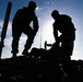 Two individuals are silhouetted against a bright blue sky, working on or near rocky terrain. They appear to be handling equipment, with a cylindrical pole or tube visible on the left side and additional equipment structures in the background. The strong backlit composition creates dramatic dark silhouettes with minimal detail visible on the subjects.