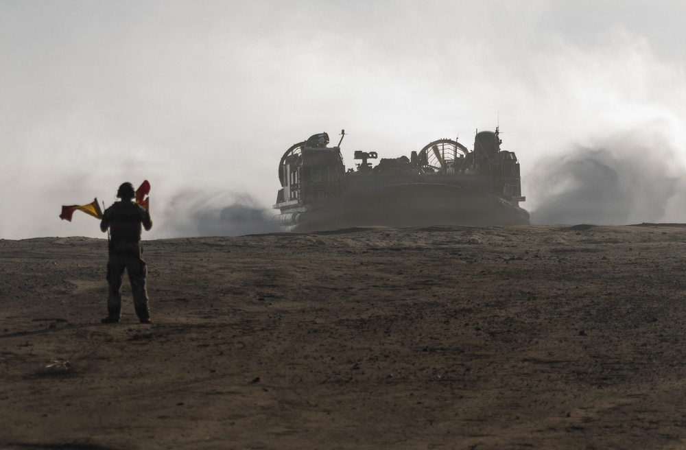 A military signalman stands in the foreground holding semaphore flags, guiding a Landing Craft Air Cushion (LCAC) hovercraft approaching across a sandy, arid terrain. The LCAC is generating significant dust and exhaust clouds as it moves forward. The scene appears to be a military amphibious landing exercise or operation.