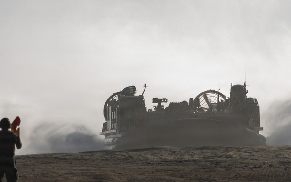 A military signalman stands in the foreground holding semaphore flags, guiding a Landing Craft Air Cushion (LCAC) hovercraft approaching across a sandy, arid terrain. The LCAC is generating significant dust and exhaust clouds as it moves forward. The scene appears to be a military amphibious landing exercise or operation.
