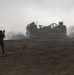 A military signalman stands in the foreground holding semaphore flags, guiding a Landing Craft Air Cushion (LCAC) hovercraft approaching across a sandy, arid terrain. The LCAC is generating significant dust and exhaust clouds as it moves forward. The scene appears to be a military amphibious landing exercise or operation.