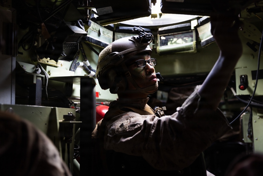 A soldier in full combat gear is positioned inside the cramped interior of a military vehicle, appearing to operate or adjust equipment overhead. The individual is wearing a tactical helmet with night vision device mount, glasses, and camouflage uniform with face paint applied. The interior is filled with various military electronics, cables, and equipment mounted on olive-green walls.