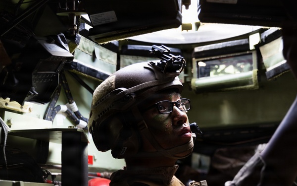 A soldier in full combat gear is positioned inside the cramped interior of a military vehicle, appearing to operate or adjust equipment overhead. The individual is wearing a tactical helmet with night vision device mount, glasses, and camouflage uniform with face paint applied. The interior is filled with various military electronics, cables, and equipment mounted on olive-green walls.