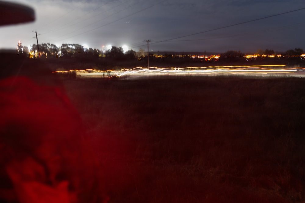 A long-exposure night photograph taken from or near a vehicle, showing light trails from passing traffic along a road running through a rural or semi-rural landscape. The extended shutter speed has captured streaks of white headlights and orange/red taillights creating wavy lines across the middle of the frame. A red-illuminated object, likely part of a vehicle interior or exterior, is visible in the foreground left.
