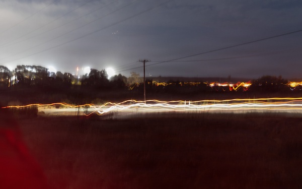 A long-exposure night photograph taken from or near a vehicle, showing light trails from passing traffic along a road running through a rural or semi-rural landscape. The extended shutter speed has captured streaks of white headlights and orange/red taillights creating wavy lines across the middle of the frame. A red-illuminated object, likely part of a vehicle interior or exterior, is visible in the foreground left.