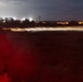 A long-exposure night photograph taken from or near a vehicle, showing light trails from passing traffic along a road running through a rural or semi-rural landscape. The extended shutter speed has captured streaks of white headlights and orange/red taillights creating wavy lines across the middle of the frame. A red-illuminated object, likely part of a vehicle interior or exterior, is visible in the foreground left.