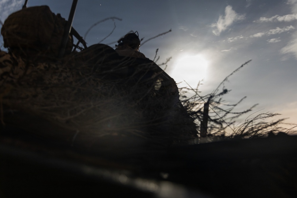 A low-angle, backlit photograph showing a figure in military camouflage gear positioned in what appears to be a field or trench environment. The image is heavily silhouetted due to the sun being directly behind the subject, creating strong contrast between the dark foreground and bright sky. Sparse, bare branches and dry vegetation are visible in the background.