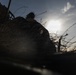 A low-angle, backlit photograph showing a figure in military camouflage gear positioned in what appears to be a field or trench environment. The image is heavily silhouetted due to the sun being directly behind the subject, creating strong contrast between the dark foreground and bright sky. Sparse, bare branches and dry vegetation are visible in the background.