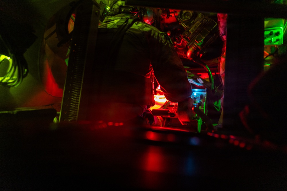 A low-light image showing what appears to be the interior of a military vehicle or aircraft cockpit illuminated by red and green instrument lighting. A person in military gear or flight suit is visible working with equipment in a confined, instrument-dense space. The scene is characteristic of night operations where red lighting is used to preserve night vision.