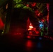 A low-light image showing what appears to be the interior of a military vehicle or aircraft cockpit illuminated by red and green instrument lighting. A person in military gear or flight suit is visible working with equipment in a confined, instrument-dense space. The scene is characteristic of night operations where red lighting is used to preserve night vision.
