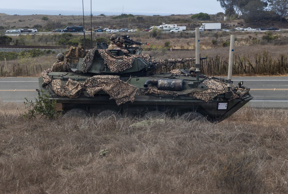A military wheeled armored vehicle, consistent with a Light Armored Vehicle (LAV), is positioned beside a road in a dry, coastal scrubland environment. The vehicle is draped with desert camouflage netting and has at least two soldiers visible on top, one of whom is aiming a rifle. Civilian vehicles and infrastructure are visible in the background, suggesting proximity to a public road or base perimeter.