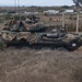 A military wheeled armored vehicle, consistent with a Light Armored Vehicle (LAV), is positioned beside a road in a dry, coastal scrubland environment. The vehicle is draped with desert camouflage netting and has at least two soldiers visible on top, one of whom is aiming a rifle. Civilian vehicles and infrastructure are visible in the background, suggesting proximity to a public road or base perimeter.