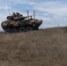 An 8-wheeled armored fighting vehicle, consistent with a Light Armored Vehicle (LAV) or similar wheeled APC, is positioned on a grassy hillside with at least one soldier visible in the open hatch. The vehicle is draped with camouflage netting and equipped with multiple antennas and a mounted weapon system. The setting appears to be a dry, open training or operational terrain under a partly cloudy sky.