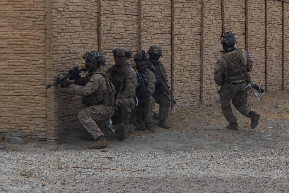 The image shows a group of military personnel in full combat gear conducting what appears to be a tactical urban operations exercise or operation. Four soldiers are stacked against a corner of a tan brick wall in a breaching formation, while a fifth soldier moves separately along the wall to the right. The environment appears to be a desert or arid urban setting.