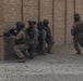 The image shows a group of military personnel in full combat gear conducting what appears to be a tactical urban operations exercise or operation. Four soldiers are stacked against a corner of a tan brick wall in a breaching formation, while a fifth soldier moves separately along the wall to the right. The environment appears to be a desert or arid urban setting.