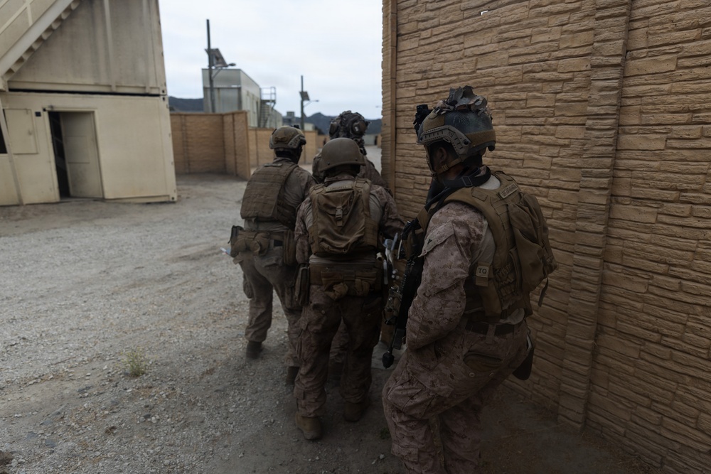 A group of military personnel in full combat gear are moving tactically along a building wall in what appears to be a training environment or military installation. The soldiers are equipped with helmets, body armor, tactical vests, and firearms, moving in a staggered formation. The setting includes modular/prefabricated structures and a gravel surface consistent with a military training facility.
