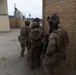 A group of military personnel in full combat gear are moving tactically along a building wall in what appears to be a training environment or military installation. The soldiers are equipped with helmets, body armor, tactical vests, and firearms, moving in a staggered formation. The setting includes modular/prefabricated structures and a gravel surface consistent with a military training facility.