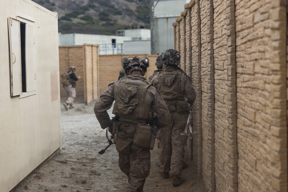 The image shows a group of military personnel in full combat gear moving through a narrow alleyway between tan/beige structures, consistent with a military training exercise or urban operations environment. The soldiers are wearing camouflage uniforms, tactical vests, helmets with night-vision goggle mounts, and carrying equipment. The setting appears to be a Military Operations in Urban Terrain (MOUT) training facility, likely in the United States based on the terrain and infrastructure visible in the background.