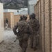 The image shows a group of military personnel in full combat gear moving through a narrow alleyway between tan/beige structures, consistent with a military training exercise or urban operations environment. The soldiers are wearing camouflage uniforms, tactical vests, helmets with night-vision goggle mounts, and carrying equipment. The setting appears to be a Military Operations in Urban Terrain (MOUT) training facility, likely in the United States based on the terrain and infrastructure visible in the background.