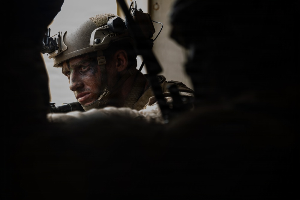 A soldier wearing tactical combat gear is visible through a gap between two silhouetted figures or objects in the foreground. The soldier has camouflage face paint applied and is wearing a ballistic helmet equipped with night-vision goggle mounts and other attachments. The image is dramatically lit, with the central subject sharply focused against a dark, shadowed composition.
