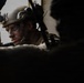 A soldier wearing tactical combat gear is visible through a gap between two silhouetted figures or objects in the foreground. The soldier has camouflage face paint applied and is wearing a ballistic helmet equipped with night-vision goggle mounts and other attachments. The image is dramatically lit, with the central subject sharply focused against a dark, shadowed composition.