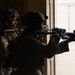 Two military personnel in tactical gear are conducting a building entry or clearing operation at a doorway. One soldier is visible in profile with camouflage face paint and a helmet with attachments, while the second is silhouetted aiming a rifle through a partially open door. The high-contrast lighting from the doorway creates a dramatic silhouette effect.