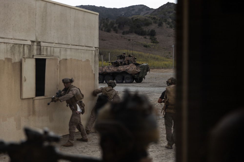 Military personnel in combat gear are conducting what appears to be a training exercise in an urban operations environment. Soldiers are moving tactically around a building structure while an armored wheeled vehicle with camouflage netting is positioned in the background. The setting appears to be a military training facility with constructed buildings and a hilly terrain backdrop.