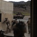 Military personnel in combat gear are conducting what appears to be a training exercise in an urban operations environment. Soldiers are moving tactically around a building structure while an armored wheeled vehicle with camouflage netting is positioned in the background. The setting appears to be a military training facility with constructed buildings and a hilly terrain backdrop.