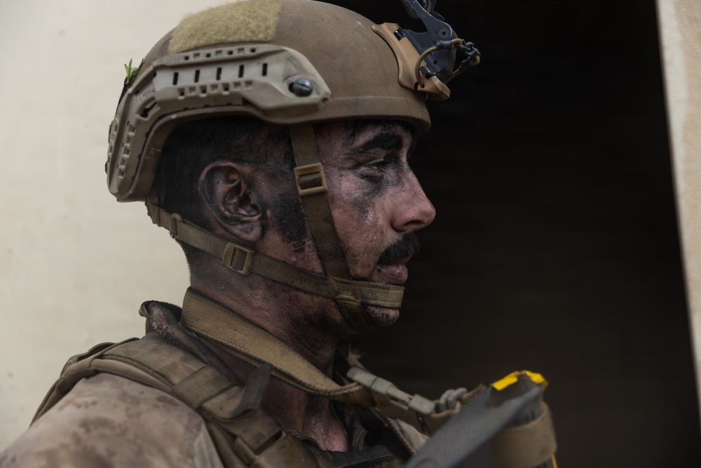 A close-up profile shot of a male soldier wearing tan/coyote brown tactical gear including a ballistic helmet with night vision mount bracket. His face is covered in dark camouflage face paint or grime, and he appears to be in a focused, alert posture. The image has a documentary or combat photography style with shallow depth of field.