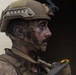 A close-up profile shot of a male soldier wearing tan/coyote brown tactical gear including a ballistic helmet with night vision mount bracket. His face is covered in dark camouflage face paint or grime, and he appears to be in a focused, alert posture. The image has a documentary or combat photography style with shallow depth of field.