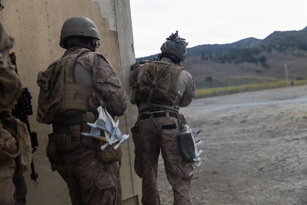 Two military personnel in full combat gear are positioned against a concrete wall, appearing to conduct a tactical exercise or training operation. Both soldiers are wearing desert camouflage uniforms, tactical vests, and helmets, and are carrying what appear to be small drone components or similar equipment attached to their gear. The setting appears to be a training range or mock urban environment with hills visible in the background.